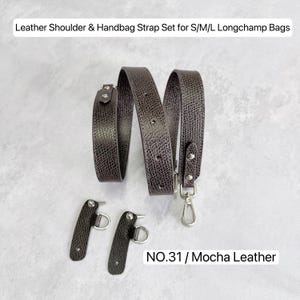 Könnte beinhalten: Ein Schulter- und Handtaschenriemen-Set aus mokkabraunem Leder mit strukturierter Oberfläche und silberfarbenen Metallbeschlägen, bestehend aus einem langen, verstellbaren Riemen mit Karabinerhaken und zwei kleineren Befestigungslaschen, beschriftet mit NO.31 / Mocha Leather.