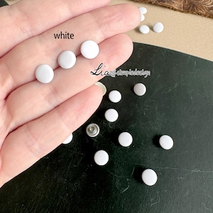May include: White round snap buttons, also known as poppers, are scattered on a dark surface. The buttons are small and have a smooth, flat surface. The text "white" is written on a hand holding some of the buttons.
