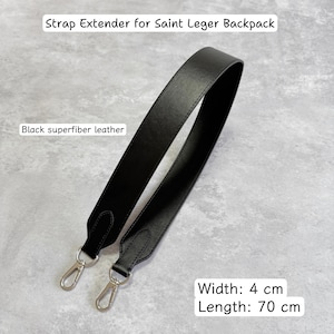 May include: Black superfiber leather strap extender for a Saint Leger backpack. The strap is 4 cm wide and 70 cm long, featuring silver-colored metal clasps.