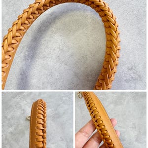 May include: Close-up of a tan leather handbag handle with a braided design. The handle is curved and appears sturdy. The leather has a smooth texture and a warm, natural colour. The handle is attached to the bag with gold-coloured hardware.