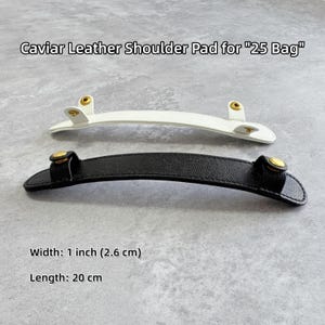 May include: Two leather shoulder pads, one white and one black, for a "25 Bag". The pads have gold-colored hardware. The width is 1 inch (2.6 cm) and the length is 20 cm. The text "Caviar Leather Shoulder Pad for "25 Bag"" is at the top.