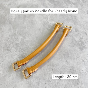 May include: Two honey-colored leather handles for a Speedy Nano bag. Each handle is 20 cm long and features gold-colored metal hardware. The text "Honey patina handle for Speedy Nano" is visible at the top of the image.