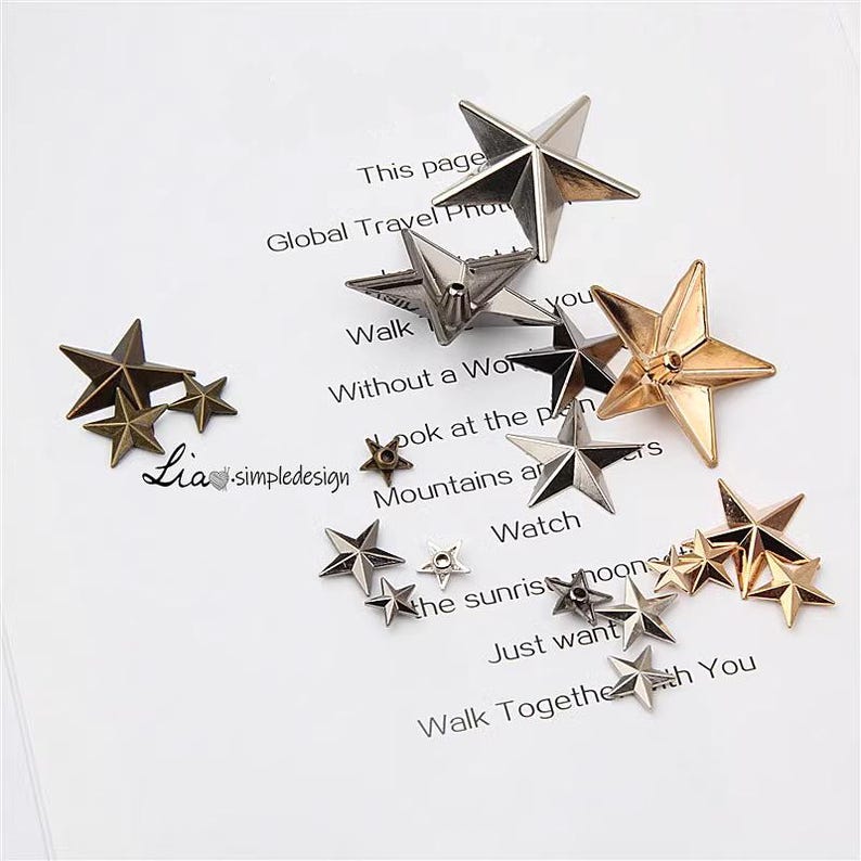 May include: Assortment of five-pointed star shaped metal embellishments in silver, gold, and bronze tones. The stars are various sizes and have a raised, textured surface.