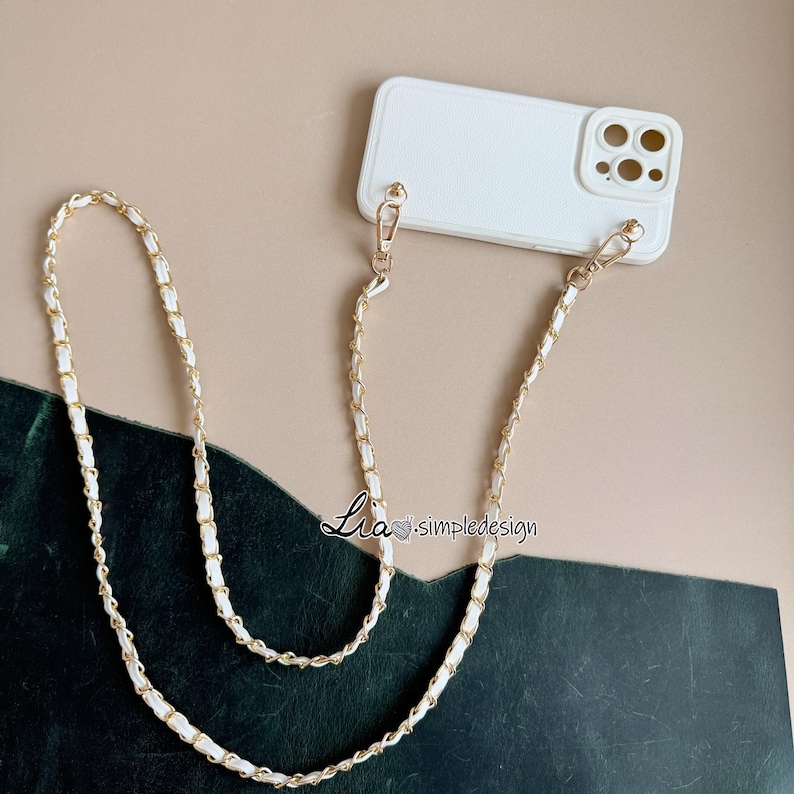 May include: White phone case with a gold chain strap. The strap is made of gold chain links interwoven with white material. The phone case is white and has a camera cutout. The text "Lia simpledesign" is visible.