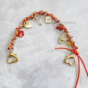 May include: An orange and gold braided strap with gold-colored heart-shaped clasps and various charms. Charms include a key, butterfly, heart, star, and a red ring. The text "Lia simpledesign" is visible.