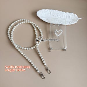 May include: A clear phone case with a pearl heart design and a white acrylic pearl strap. The strap is 120 cm long and features silver-toned accents. The text "Lia simpledesign" is visible on the case.
