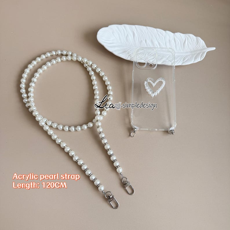 May include: A clear phone case with a pearl heart design and a white acrylic pearl strap. The strap is 120 cm long and features silver-toned accents. The text "Lia simpledesign" is visible on the case.