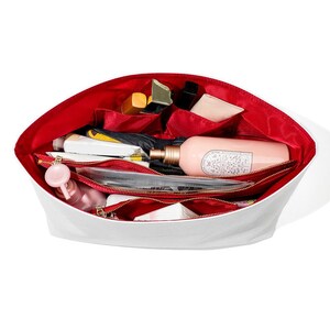 May include: A white cosmetic bag with a red interior, filled with various beauty products. The bag has multiple compartments and zippered pockets, and contains items like lipstick, lotion, and a small spray bottle.