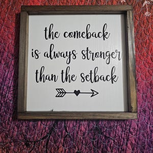 May include: A wooden framed sign with a white background and black lettering. The sign reads "the comeback is always stronger than the setback" with an arrow and heart in the center.