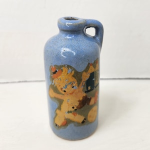 May include: A small, blue ceramic jug with a handle. The jug features a colorful illustration of a bear-like character and a house. The jug is likely vintage and suitable for decorative purposes.