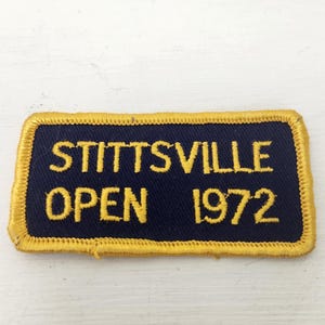 May include: A rectangular patch with the words "STITTSVILLE OPEN 1972" embroidered in yellow on a navy blue background. The patch has a gold border and appears to be vintage.