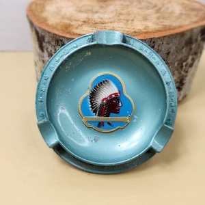 May include: A blue metal ashtray with a Native American chief design. The ashtray has a raised rim and a small indentation in the center. The chief is wearing a feathered headdress and has a red face.