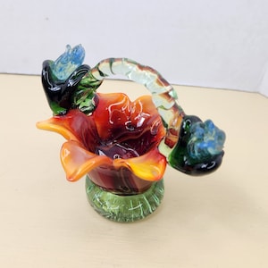 May include: A small glass basket with a flower-shaped base and a twisted handle. The basket is decorated with red, orange, yellow, green, and blue glass.