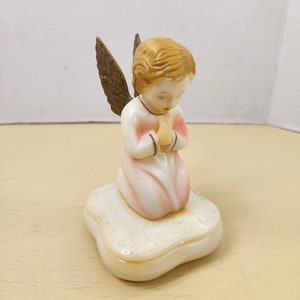 May include: A small, white porcelain figurine of a praying angel with gold wings. The angel is kneeling on a white, heart-shaped cushion.