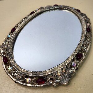 May include: A gold-toned oval mirror with a decorative frame. The frame is adorned with red and iridescent rhinestones.