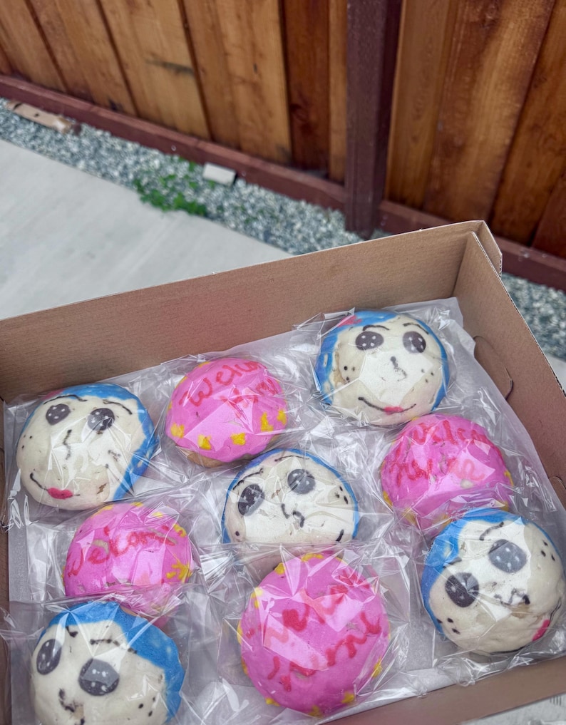 May include: A cardboard box filled with individually wrapped, round baked goods. Some have blue frosting with a face design, others are pink with text. The baked goods are arranged in rows.