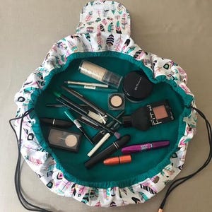 May include: A round, drawstring make-up bag featuring a white exterior with a colourful feather pattern in pink, teal, and black. The interior is a solid teal fabric, currently filled with various cosmetic items including lipsticks, brushes, and compacts.