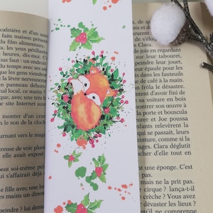 Peut inclure: Marque-page blanc avec une illustration à l'aquarelle de deux renards orange nichés ensemble, entourés d'une couronne de feuilles vertes et de baies rouges. Des éclaboussures de peinture verte et orange décorent le marque-page.