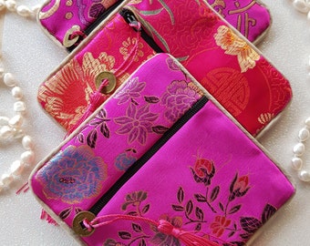 May include: Three pink fabric pouches with gold accents and tassels. The pouches are decorated with floral patterns and have zips.