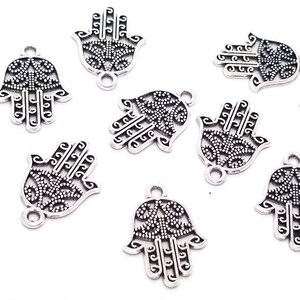 Puede incluir: Nueve dijes de metal plateado con forma de mano de Hamsa, con intrincados recortes de filigrana y un peque&ntilde;o lazo en la parte inferior. Una moneda de diez centavos de EE. UU. a la izquierda sirve de escala, midiendo unos 1,9 cm de alto.
