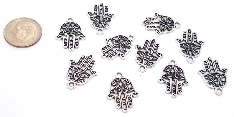 Puede incluir: Nueve dijes de metal plateado con forma de mano de Hamsa, con intrincados recortes de filigrana y un peque&ntilde;o lazo en la parte inferior. Una moneda de diez centavos de EE. UU. a la izquierda sirve de escala, midiendo unos 1,9 cm de alto.