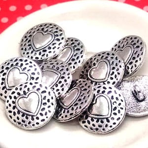 May include: A group of silver-toned metal buttons with a heart design. Each button has a raised heart in the center with a textured background.