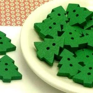 May include: A collection of small, flat, green wooden buttons shaped like Christmas trees. Each button features two holes in the center for attachment. Several buttons are clustered together on a white plate, while three individual buttons are placed separately on the surface.