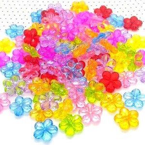 May include: A collection of colorful, translucent flower-shaped beads in various shades of pink, blue, yellow, orange, and red. The beads are scattered on a white surface, creating a vibrant and playful display. These beads are ideal for jewelry making and craft projects.