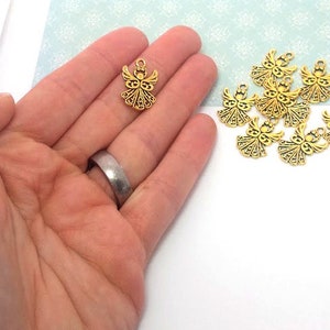 May include: A close-up of a hand holding a gold angel charm. There are several other gold angel charms scattered on a white surface.