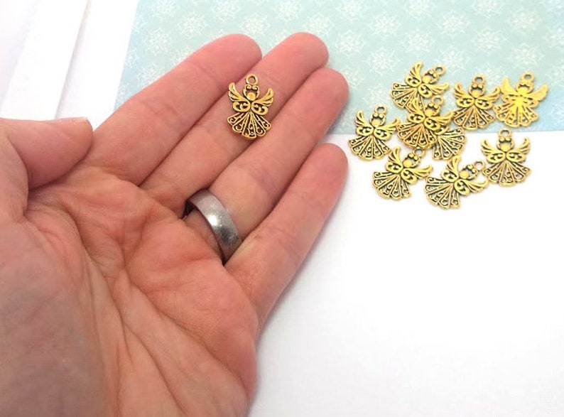 May include: A close-up of a hand holding a gold angel charm. There are several other gold angel charms scattered on a white surface.