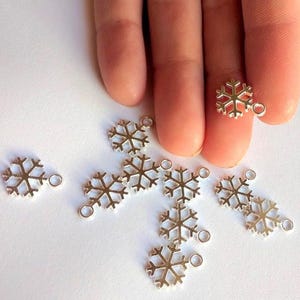 May include: A collection of small, silver-toned metal snowflake charms, each featuring a six-pointed geometric design and a single circular jump ring at the top for attachment. The charms are arranged in a scattered pattern on a plain surface.