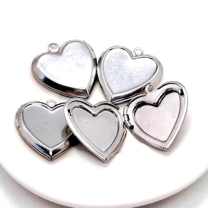 May include: Five silver heart-shaped lockets, some open, some closed, with a small loop at the top for a chain. The lockets are on a white surface. The heart-shaped lockets are a romantic jewelry item.
