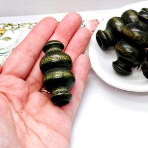 May include: Five dark green wooden beads with a textured surface. The beads are shaped like small vases with a rounded bottom and a narrow neck.
