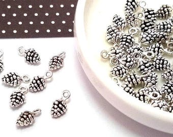 May include: Silver-colored pine cone charms with a loop for jewelry making. Some charms are scattered on a white surface, while others are in a white dish. A brown and white polka dot pattern is in the background.