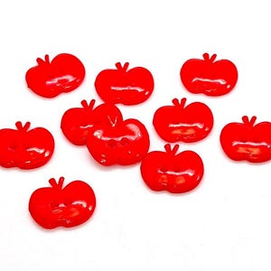 May include: A collection of ten bright red, apple-shaped buttons. Each button features a smooth, glossy finish with two small holes in the centre for attachment. The buttons are arranged in a scattered pattern against a plain white background.