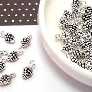 May include: Silver tone metal pine cone charms with a loop for hanging. The charms are scattered on a white surface.