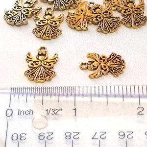 May include: Nine gold angel charms with intricate details, measuring approximately 1/32 inch in size. The charms are arranged on a white surface with a ruler for scale.