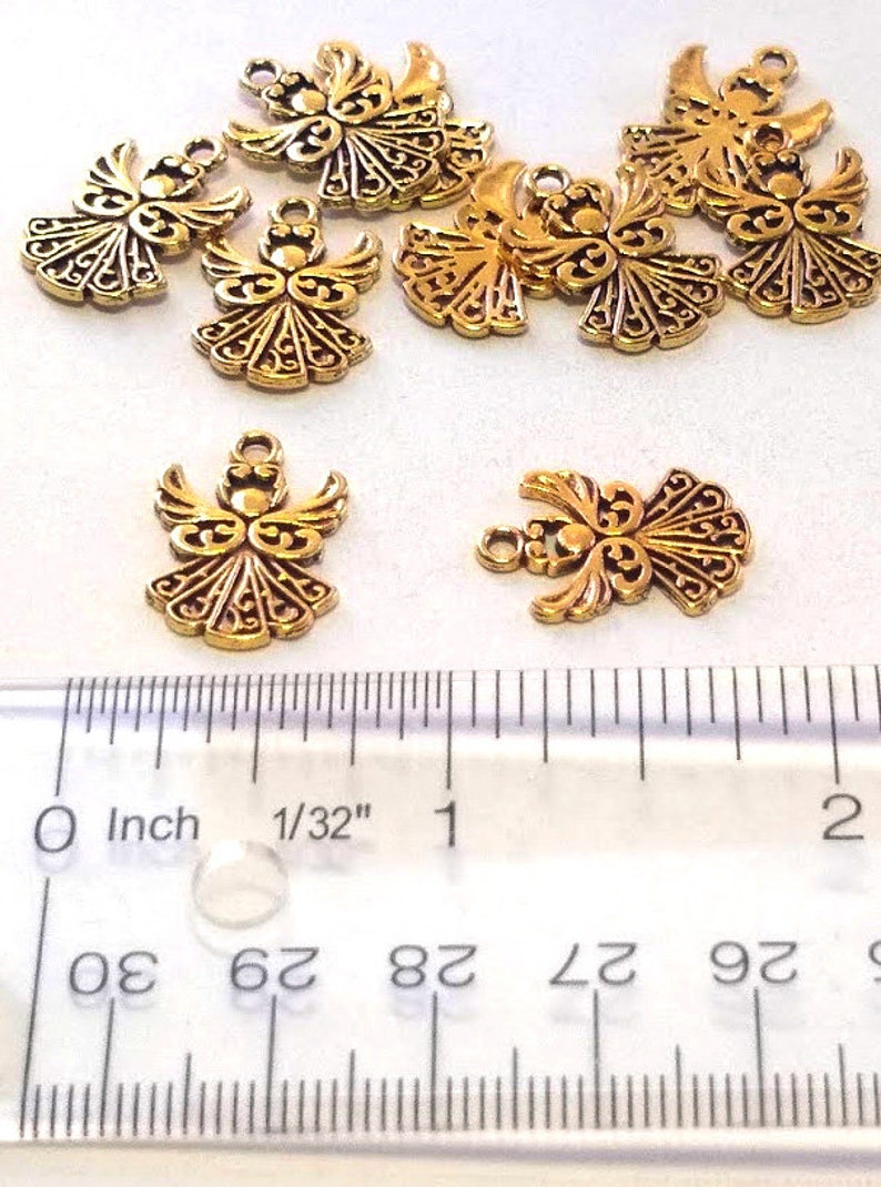 May include: Nine gold angel charms with intricate details, measuring approximately 1/32 inch in size. The charms are arranged on a white surface with a ruler for scale.