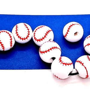May include: A collection of nine spherical white beads, each featuring a red stitched pattern resembling a baseball. The beads are strung on a thin black cord, arranged in a loose, curved formation across the frame to display the design.