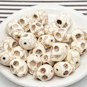May include: A collection of off-white skull-shaped beads with brown veining, displayed on a white ceramic plate. The beads have holes for stringing and are suitable for jewelry making or craft projects. The skulls are approximately 1 inch in size.