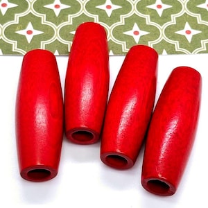 May include: Four red wooden beads, each with a hole in the center, are arranged in a row on a white surface. The beads are oval-shaped and have a smooth, polished finish.