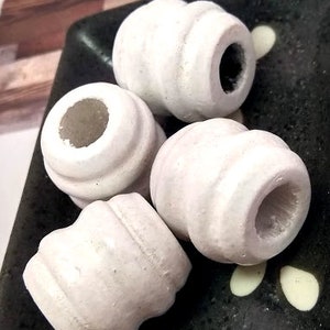 May include: Four white, cylindrical beads with a ridged design are arranged on a dark, textured surface. Each bead has a large, central hole. The beads are a matte white color.