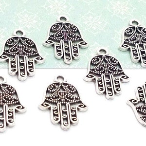 Puede incluir: Un conjunto de nueve dijes de metal plateado con forma de mano de Hamsa, con intrincados patrones de filigrana y puntos. Cada dije incluye una peque&ntilde;a anilla en la parte superior para colgar. Una moneda de diez centavos de EE. UU. sirve de referencia.