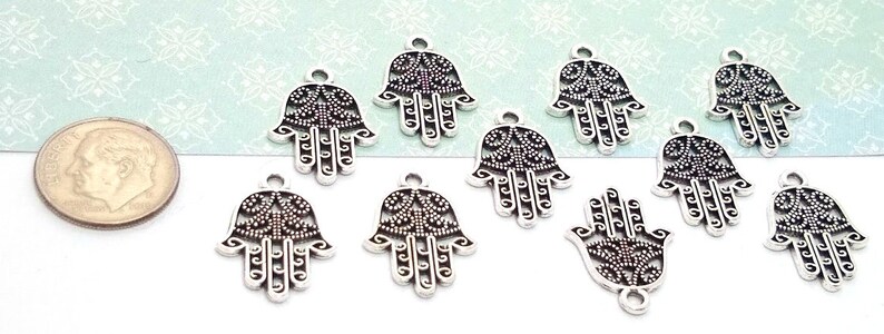 Puede incluir: Un conjunto de nueve dijes de metal plateado con forma de mano de Hamsa, con intrincados patrones de filigrana y puntos. Cada dije incluye una peque&ntilde;a anilla en la parte superior para colgar. Una moneda de diez centavos de EE. UU. sirve de referencia.