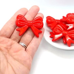 May include: Five red plastic bow-shaped embellishments. The bows are glossy and have a slightly 3D appearance.