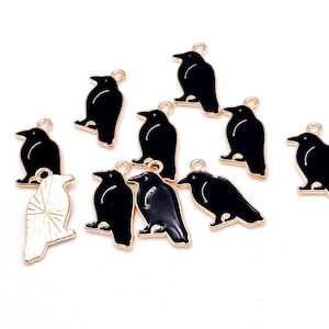 May include: A collection of black raven charms with gold-colored trim and a loop for attaching to jewelry or other crafts. One charm is white with gold-colored accents. The charms are on a white background.