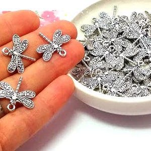 May include: Silver-colored dragonfly charms. Three charms are held in a hand, with a ring on the finger. A small white bowl is filled with more dragonfly charms. The charms have detailed wing patterns and a loop for attaching to jewelry.