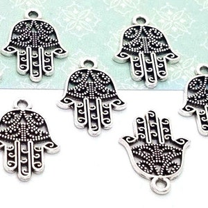 Puede incluir: Un conjunto de nueve dijes de mano de Hamsa en metal plateado. Cada dije presenta un intrincado dise&ntilde;o de filigrana calada con un acabado oscuro oxidado en las &aacute;reas hundidas y un peque&ntilde;o lazo circular en la parte superior para colgar.
