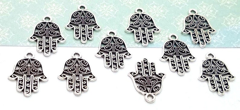 Puede incluir: Un conjunto de nueve dijes de mano de Hamsa en metal plateado. Cada dije presenta un intrincado dise&ntilde;o de filigrana calada con un acabado oscuro oxidado en las &aacute;reas hundidas y un peque&ntilde;o lazo circular en la parte superior para colgar.