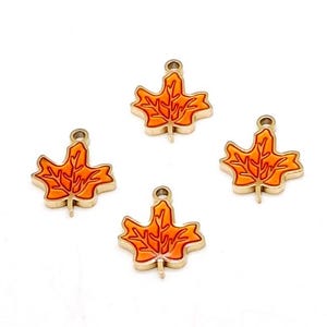 May include: Four gold-toned metal charms shaped like maple leaves, each featuring a vibrant orange enamel inlay with raised gold vein details and a small circular loop at the top for attachment, arranged in a scattered pattern on a white background.